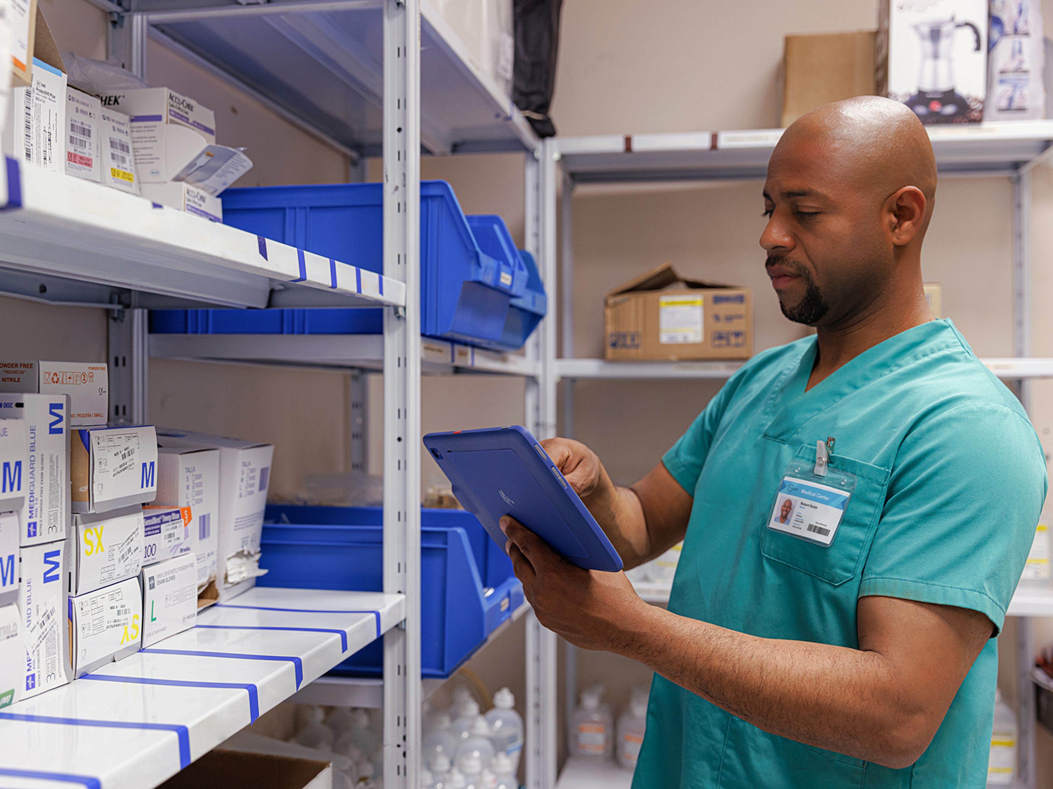 A healthcare professional is in a medical supply warehouse, holding a tablet and searching for information regarding the supplies.