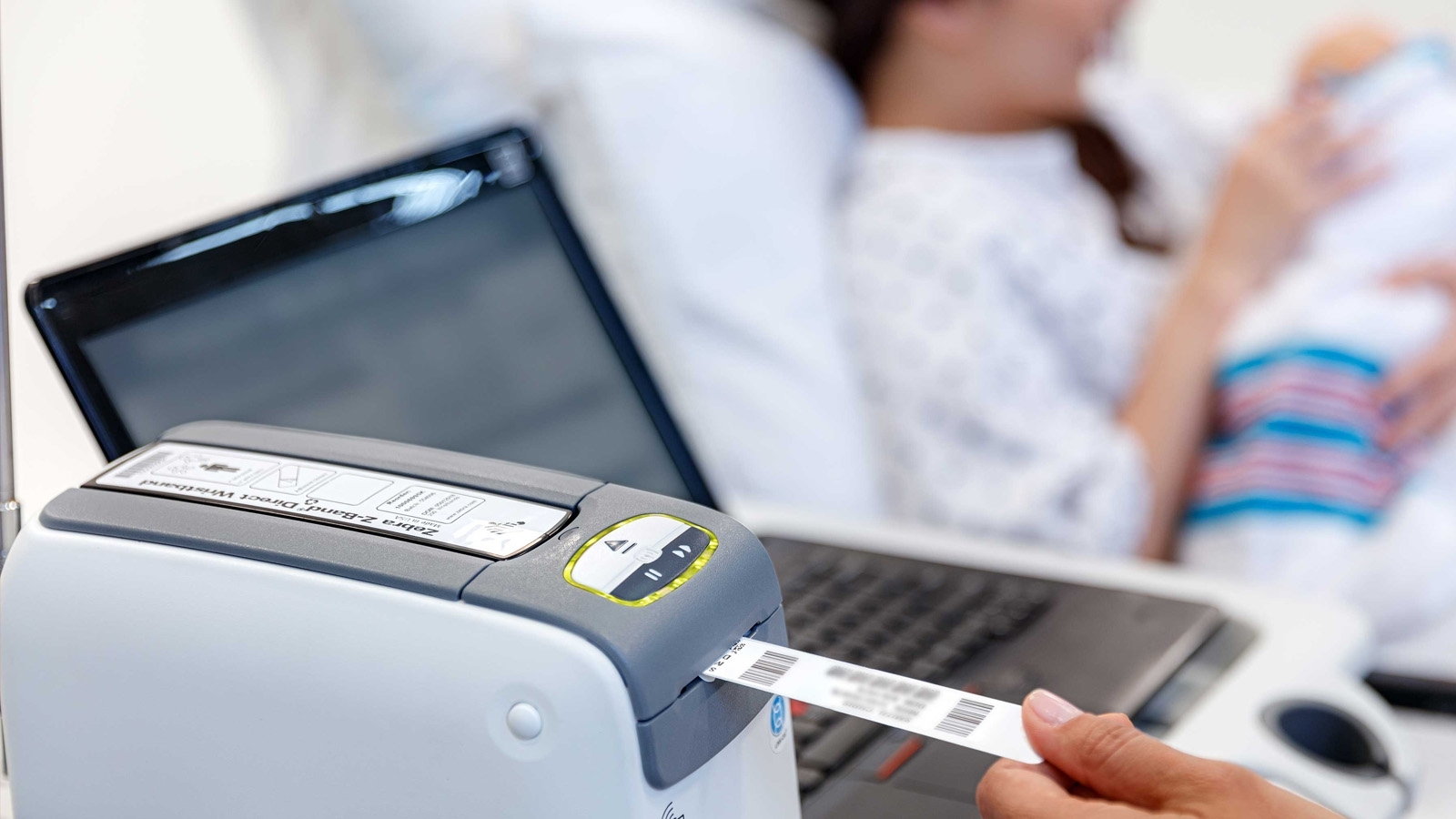 The Zebra Patient Identification wristband is being printed using a Zebra printer next to a patient holding a newborn while lying on a bed. 