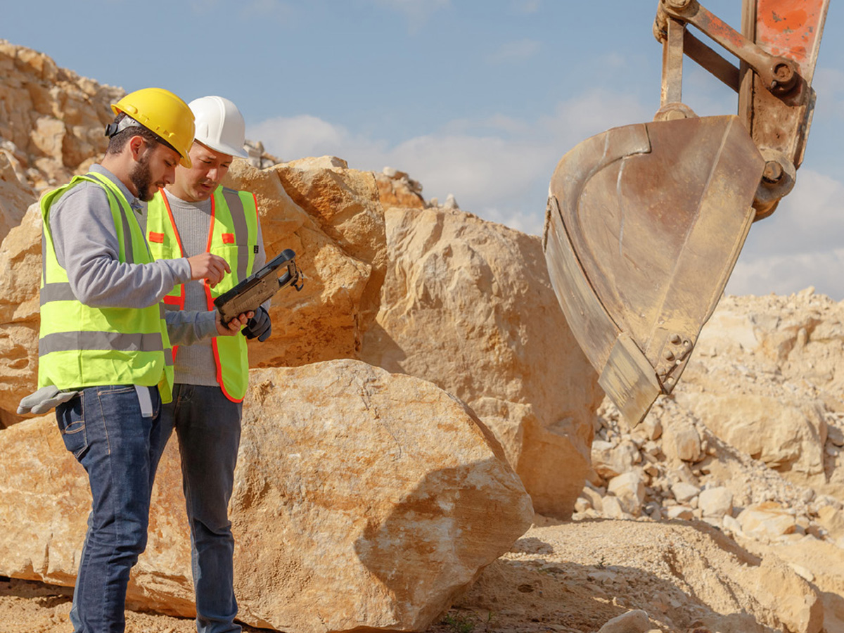 Two workers together with one holding a zebra tablet standing in front of a digger bucket