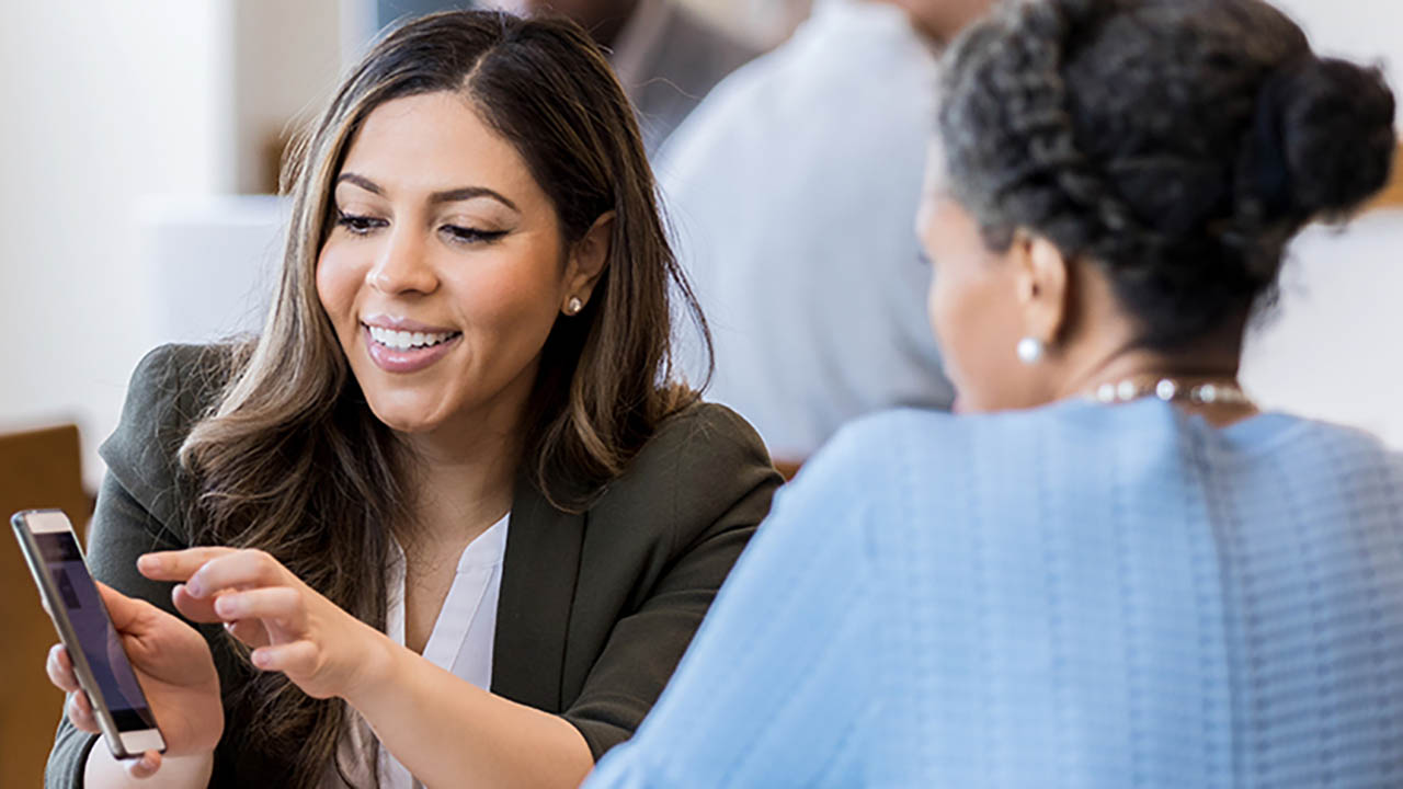 Mid adult female bank employee discusses a mobile banking app's features with a mature female client.