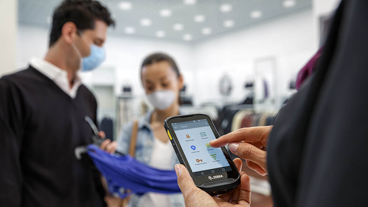 A department store associate uses a handheld mobile computer to look up something while keeping a safe distant from customers.