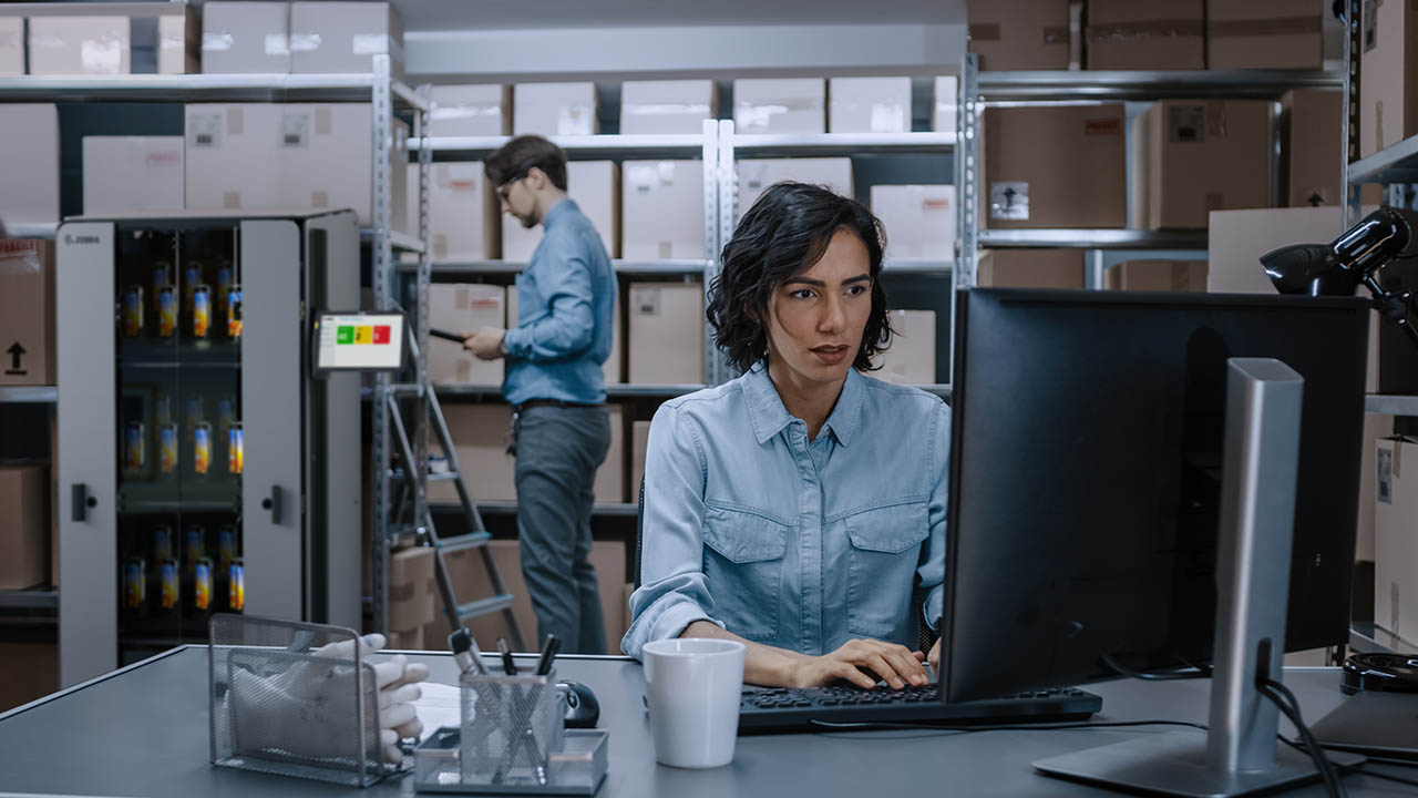 Warehouse Female Inventory Manager Works on a Computer while Sitting at Her Desk, In the Background Male Worker Uses Digital Tablet Computer To Check Shelf for a Delivery Package.